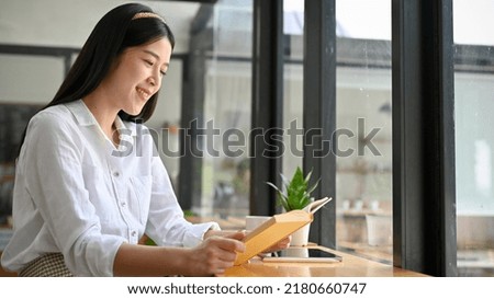Pretty and beautiful young Asian woman or female college student relaxes sitting in the coffee shop and enjoy reading her favourite book.