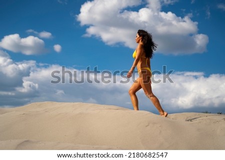 Beautiful young woman in yellow swimsuit walking on sand, copy space sky background 