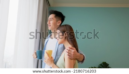 a happy asian couple is hugging and watching outside the window together at their home they are enjoying their tea simultaneously
