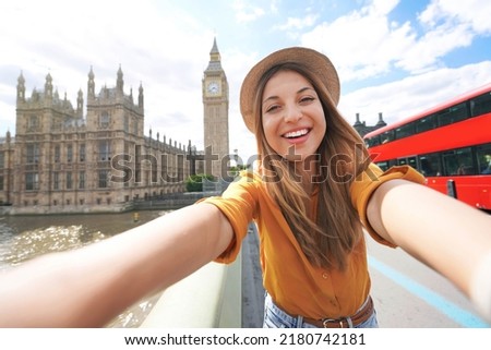 Smiling tourist girl taking self portrait in London, UK. Selfie photo of happy woman traveling in London with Big Ben tower, Westminster palace and double decker red bus on summer sunny day.
