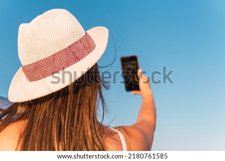 Close up shot of young brunette woman in white hat taking a selfie holding phone with one hand sitting in the window of her parked camper van. Van life concept.