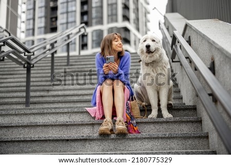 Cheerful woman sits with her cute adorable dog on staircase and using phone at modern residential area. Spending leisure time and friendship with pets concept