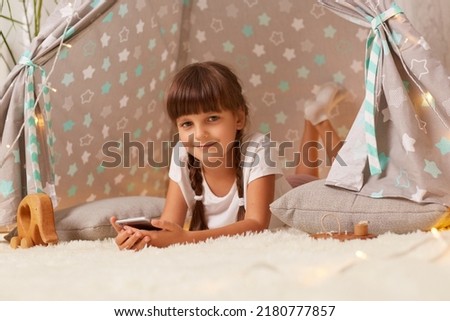 Portrait of delighted little girl with pigtails wearing white t shirt posing in wigwam at home and holding cell phone, looking at camera with positive optimistic facial expression.