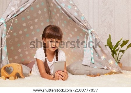Image of cute dark haired little girl with pigtails wearing white t shirt posing in wigwam at home, watching cartoons, looking at smart phone with concentrated impressed expression.