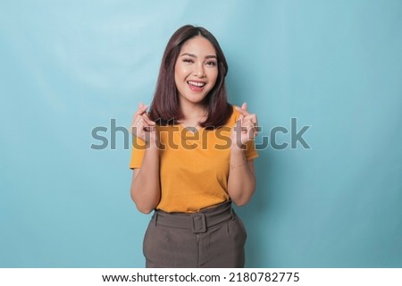 Attractive young Asian woman feels happy and romantic shapes heart gesture expresses tender feelings wears casual yellow t-shirt against blue background. People affection and care concept Royalty-Free Stock Photo #2180782775