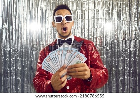 Happy wealthy ethnic winner man in shiny suit, bowtie and cool glasses looks at paper money bills bunch with surprised wow face expression. TV game show host guy in foil fringe studio shows prize cash