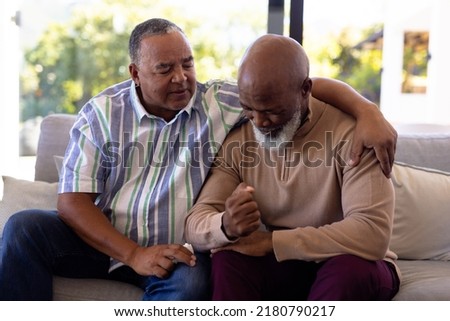 Multiracial senior man consoling male friend crying while sitting on sofa in nursing home. Sadness, togetherness, comfort, unaltered, emotional stress, support, assisted living, retirement concept. Royalty-Free Stock Photo #2180790217