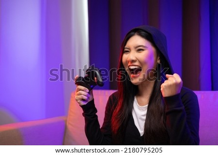 Excited and shocked face of Asian female gamer with hoodie holding joystick playing video game online sitting on sofa at living room. Chinese pro gamer streaming online social playing game very fun