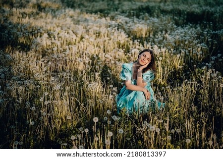Attractive woman sitting on ripened dandelion lawn in park. Girl in retro turquoise dress enjoying summer in countryside. Wishing, joy concept. Springtime, aesthetic portrait