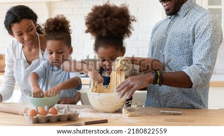 Happy emotional little african american girl making dough for homemade pastry with joyful brother and smiling biracial parents, standing together at wooden countertop in modern kitchen, family hobby.