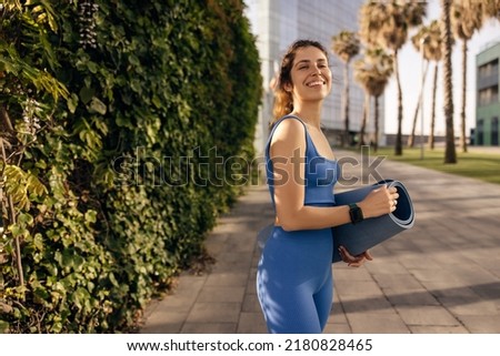 Joyful young caucasian girl smiles broadly, holds yoga mat outdoors in her hand. Brown-haired woman wears blue clothes for sports. Workout concept