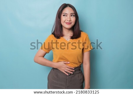 Pleased cheerful Asian woman keeps hand on belly feels full after delicious dinner dressed casually stands thoughtful against blue background. Royalty-Free Stock Photo #2180833125