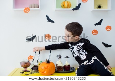 Toddler caucasian boy in skeleton costume sits on the table with Halloween food and decorations. Halloween party at home