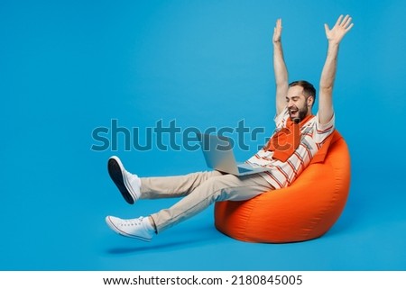 Full body young excited fun cool man 20s in orange striped t-shirt sit in bag chair hold use work on laptop pc computer with overstretched hands legs isolated on plain blue background studio portrait.