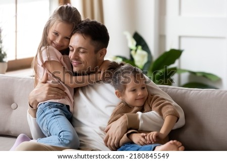 Pleased dad sit on couch with kids, after long separation missed his two little 7s daughters, closing his eyes, hugs them enjoying moment. Family bond and love, Happy Father Day celebration concept Royalty-Free Stock Photo #2180845549