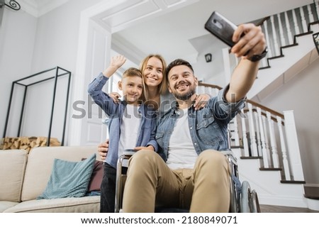 Smiling family standing near father with special needs and looking at smartphone camera during online video call. School age son waving hand while dad in wheelchair holding phone.