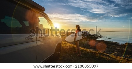 Surfergirl walking near her mini van and looking on the ocean at summer sunset  with a surfboard on her side - selective focus Royalty-Free Stock Photo #2180855097