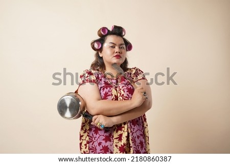 Asian Crazy Housewife. Angry Woman Raising Frying Pan Threatening  Looking At Camera Standing Over creame Studio Background. Furious Wife Shouting Tired Of Cooking Concept Royalty-Free Stock Photo #2180860387