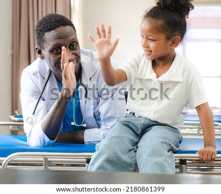 Child patient sitting on hospital bed with African doctor for medical care. Smiling little girl happy healthy after professional checkup at clinic. Practitioner, pediatrician checking, examining kid Royalty-Free Stock Photo #2180861399