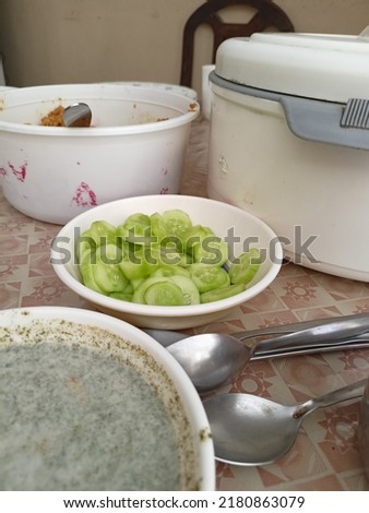 This picture contain cucumber in a bowl, some spoons, hot pot, and chutney. This is a dinner picture that looks interesting and beautiful.