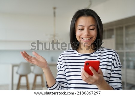 Happy girl is excited with mobile phone using. Conceptual portrait of young emotional woman holding smartphone. Pretty hispanic girl in striped casual shirt at home. Advertising banner mockup.