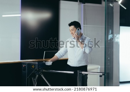 The worker enters the office, opens the door with an electronic key with a card, an Asian man smiles and talks on the phone Royalty-Free Stock Photo #2180876971