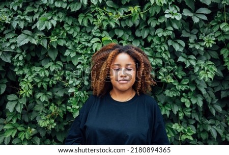 Young positive african woman smiling outside with a ivy plant in the background  at street of city wearing on black blouse and blue jeans. Calm and nature in the city concept