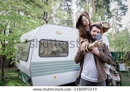 Male giving piggyback ride to girlfriend, having fun and taking a selfie. Happy young caucasian couple traveling in travel van. Romantic atmosphere of relaxation. Road trip around country for weekend
