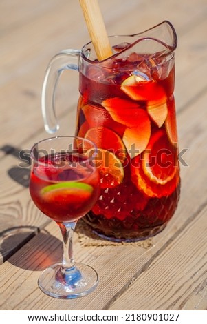 Red wine sangria decanter with apple, orange, lemon, lime wedges, raspberries, grapes and ice. There is a glass of ice, raspberries and sangria nearby. Stands on a wooden background illuminated by sun Royalty-Free Stock Photo #2180901027