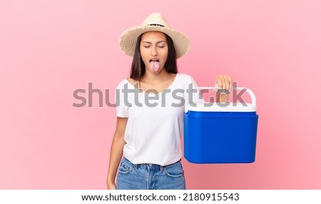 pretty hispanic woman feeling disgusted and irritated and tongue out and holding a portable refrigerator