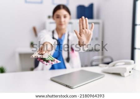 Young hispanic doctor woman wearing doctor uniform holding prescription pills at clinic with open hand doing stop sign with serious and confident expression, defense gesture 