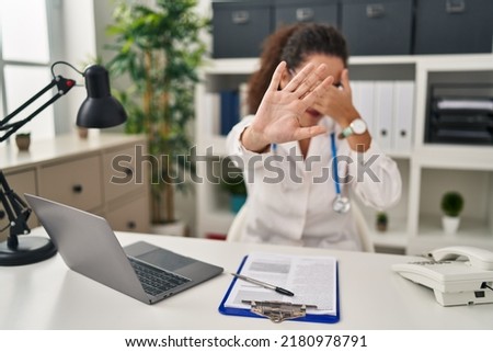 Young hispanic woman wearing doctor uniform and stethoscope covering eyes with hands and doing stop gesture with sad and fear expression. embarrassed and negative concept. 
