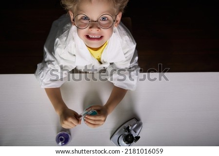 Picture from above of a smiling boy with glasses at a table with test tubes in his hands and a microscope on a black background. A child in a chemistry lesson with real chemical reagents in his hands