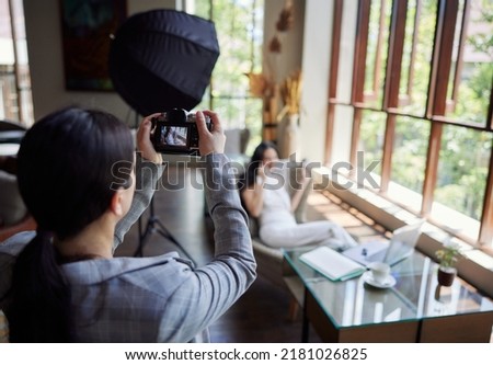 Professional female photographer takings pictures of businesswoman sitting in chair and talking on the phone. Behind the scene in photography industry. Filming stock photography content