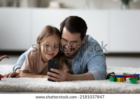Cute girl lying on carpet with dad watch new vlog on cellphone, use smartphone for leisure fun enjoy cool mobile application smiling resting on floor in playroom. Modern tech, parental control concept