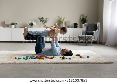 Loving dad lying on warm floor with underfloor heat system play with 6s daughter, lifts on arms cute kid, girl spread arms imagining flying in air. Family vacation, fun, active family playtime concept Royalty-Free Stock Photo #2181037853