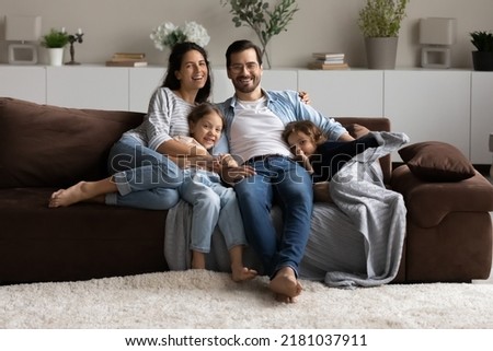 Happy couple and kids sit on sofa smiling look at camera. Preschool 5s girl, little 4s boy and parents relax together on couch posing photographing, capture moment. Well-being family portrait concept Royalty-Free Stock Photo #2181037911