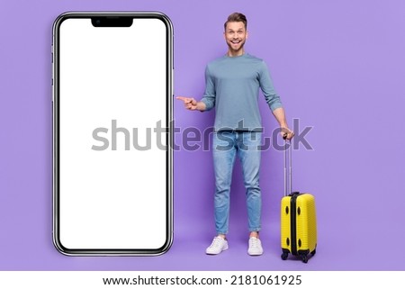 Photo of positive excited guy wear grey shirt holding suitcase pointing modern device empty space isolated violet color background