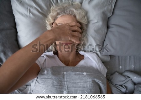 Peaceful sleepy senior woman awaking in her bed, rubbing face, covering eyes with hand from disturbing light. Elderly mature lady suffering from insomnia, sleep disorder or deprivation. Top view Royalty-Free Stock Photo #2181066565