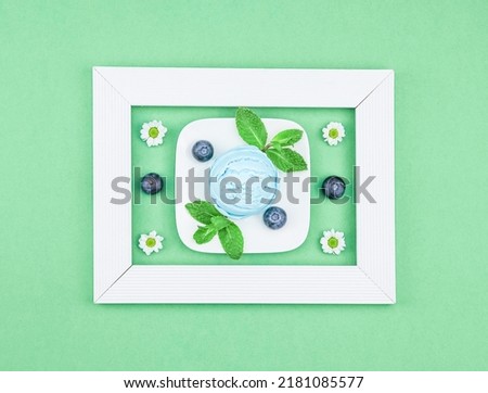 One scoop of blueberry ice cream in a square saucer with fresh berries, mint leaves and chamomile flowers in a white wooden frame on a green mint background, flat lay close-up. The concept of ice crea