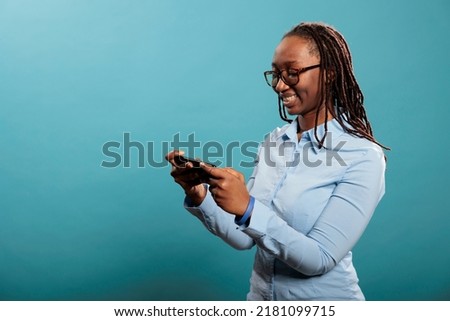 Confident joyful young adult person with modern cellphone watching video content. Beautiful happy woman with touchscreen smartphone browsing internet webpages while standing on blue background.