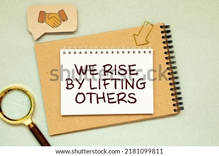 Motivation concept.Text WE RISE BY LIFTING OTHERS writing on notebook with glasses,pencil and green plant on a wooden background.