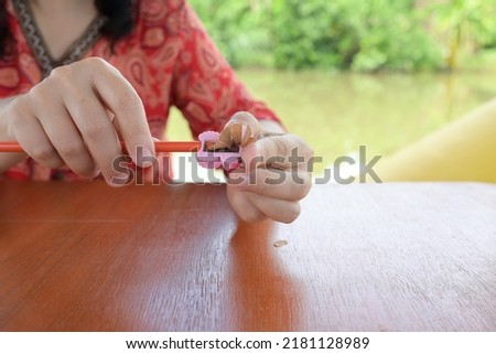 Female hands sharpens a orange pencil with a pink sharpener on wooden table.  Wooden pencil shavings come out of sharpener. Prepare a sharp pencil for convenient use. Royalty-Free Stock Photo #2181128989