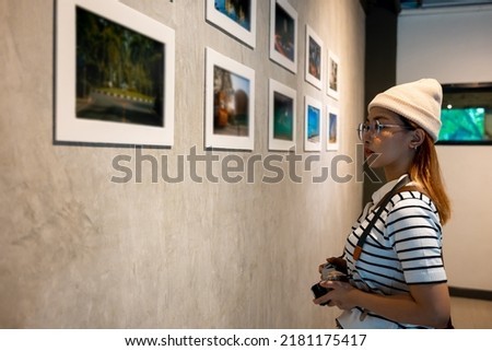 Photographer visit at photo frame to leaning against at show exhibit artwork gallery, Asian woman hold camera at picture art gallery collection in front framed paintings pictures on wall and looking