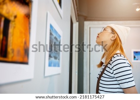 Asian woman standing she looking art gallery in front of colorful framed paintings pictures on white wall, people watch at photo frame to leaning against at show exhibition artwork gallery, Side view