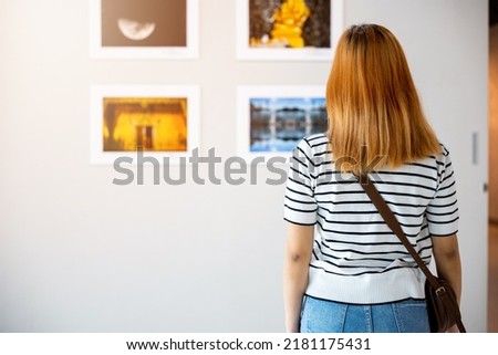 Asian woman standing she looking art gallery in front of colorful framed paintings pictures on white wall, young female watch at photo frame to leaning against at show exhibition gallery, Back view Royalty-Free Stock Photo #2181175431