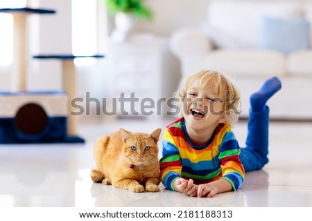Child playing with cat at home. Kids and pets. Little boy feeding and petting cute ginger color cat. Cats tree and scratcher in living room interior. Children play and feed kitten. Home animals. Royalty-Free Stock Photo #2181188313