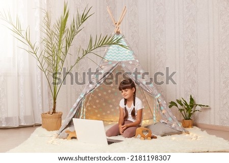 Horizontal shot of charming little girl sitting in wigwam and watching cartoons on laptop, having positive facial expression, looking at notebook screen, enjoying interesting video.