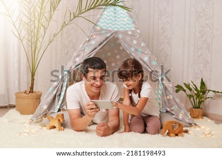 Indoor shot of happy family spending time at home, father with his daughter watching cartoons or funny video, expressing positive emotions while posing in peetee tent.