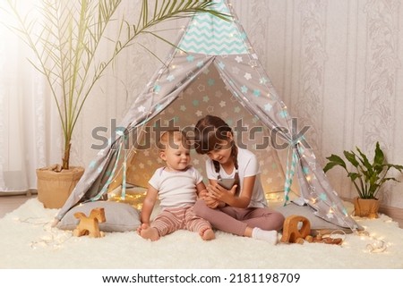 Portrait of two little girls sitting at home in teepee tent and playing on the phone, looking at device screen, watching cartoons, enjoying leisure time together.